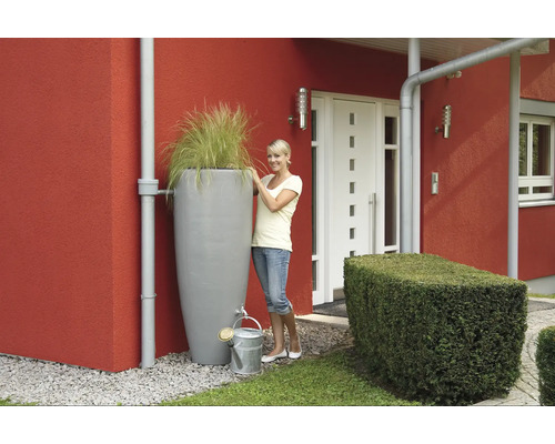 Szene mit Regenwassertank, Bepflanzung und Gießkanne vor einem Haus.