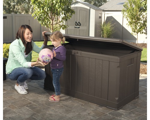 Braune Gartenbox aus Kunststoff mit geöffnetem Deckel im Garten mit Frau und Kind