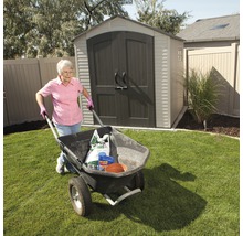 Eine Frau schiebt eine Schubkarre mit Gartengeräten und -materialien vor einem Gartenschuppen.