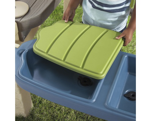 Detailansicht eines Kindes, das einen grünen Deckel auf eine blaue Wasserspielküche legt.