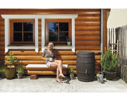 Frau sitzt auf einer Bank vor einem Holzhaus mit Regenfass