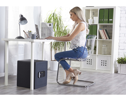 Eine Frau arbeitet an einem Schreibtisch mit einer Schreibtischlampe, einem Laptop und einem Safe im Büroraum.