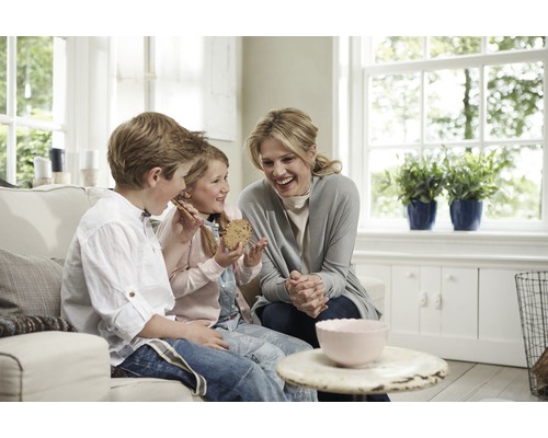 Eine Mutter und ihre Kinder sitzen auf einem Sofa und essen Kekse in einem hellen Wohnzimmer.