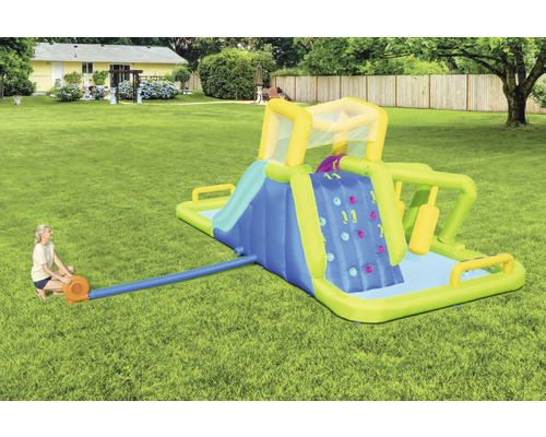 Aufblasbarer Wasserpark mit Rutsche, Kletterwand und Pool im Garten, eine Frau pumpt Luft hinein.