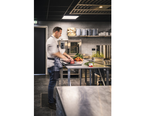 Ein Koch bereitet in einer professionellen Küche Gemüse und Fleisch auf einem Edelstahltisch zu.