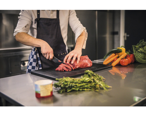 Eine Person schneidet Fleisch auf einem Schneidebrett mit Gemüse im Hintergrund.