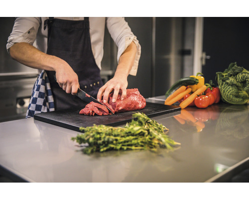 Eine Person schneidet Fleisch auf einem Schneidebrett, umgeben von Gemüse und Kräutern.