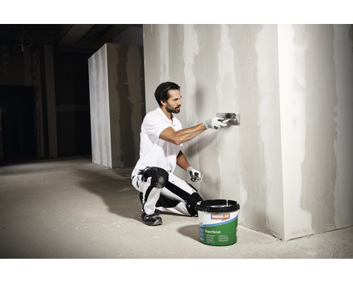 Ein Mann glättet eine Wand mit einer Spachtel neben einem Eimer Modulan Superfinish Spachtelmasse.