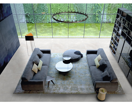 Heller Wohnraum mit Fliesenboden, Sofas, Tisch und Bücherregal vor einem großen Fenster.