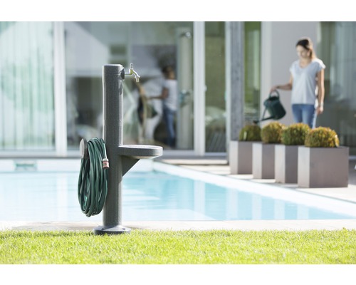Gartenszene mit Wasserzapfsäule, Wasserschlauch und Frau mit Gießkanne