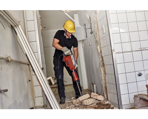 Ein Mann mit Schutzhelm und Schutzbrille arbeitet mit einem Abbruchhammer in einem gefliesten Raum.