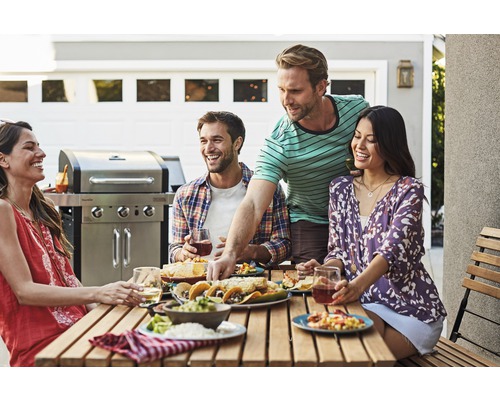 Gruppe von Freunden genießt eine Mahlzeit im Freien mit einem Grill im Hintergrund.