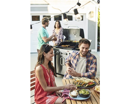 Eine Gruppe von Menschen grillt und isst im Freien, wobei ein Grill, Tacos und Guacamole auf dem Tisch stehen.