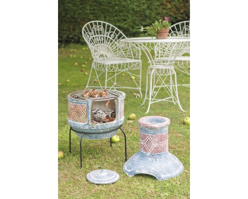 Gartenszene mit Terrakotta-Grillkamin, Grillgut, Grillrost und weißer Gartengarnitur auf einer Rasenfläche