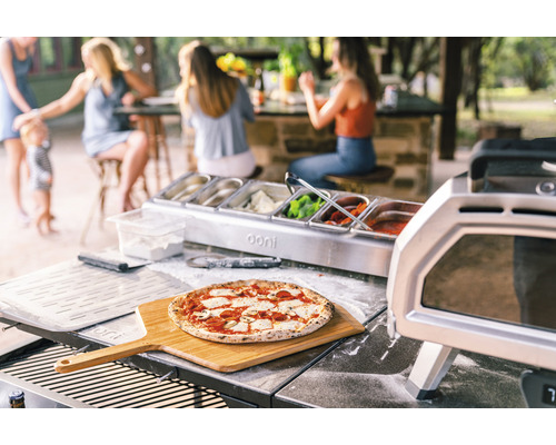 Szene im Freien mit zubereiteter Pizza auf einem Holzschieber, Ooni Pizzaofen und Zutatenbehälter