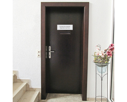 Eine dunkle Tür mit Briefkasten und Hinweisschild neben einer Treppe und einer Blumenvase.