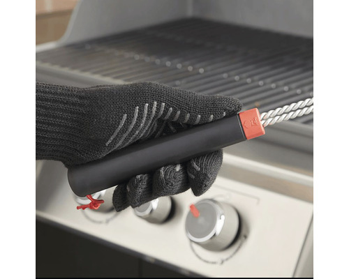 Eine Person mit Grillhandschuhen hält eine Grillbürste vor einem Grill.