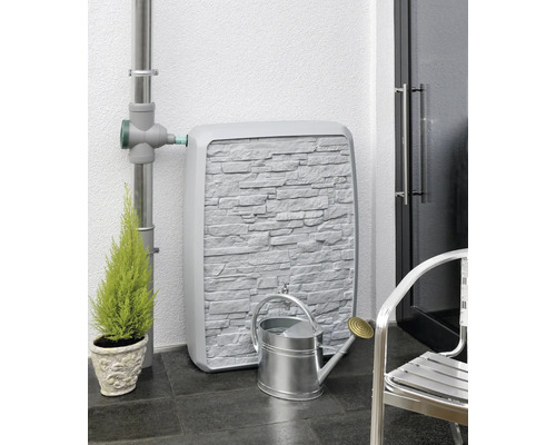 Regentonne mit Steinwandoptik, verbunden mit einem Fallrohr, daneben eine Gießkanne und eine Pflanze.