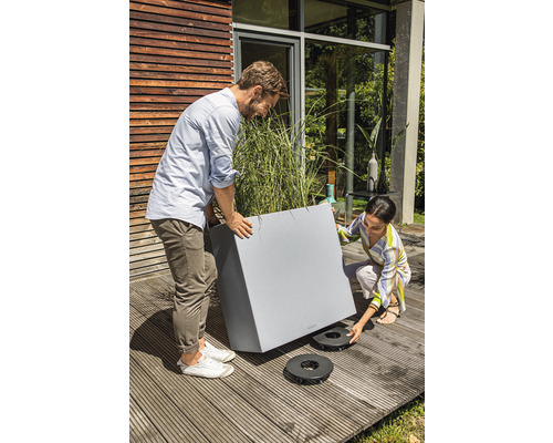 Personen platzieren einen grauen Pflanzkasten mit Pflanzen auf Pflanzenrollern auf einer Holzterrasse.