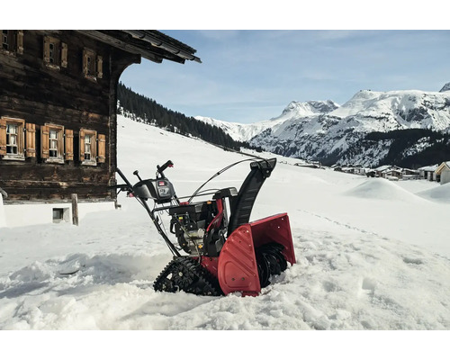 Eine Schneefräse steht im Schnee vor einem Haus in einer Winterlandschaft.