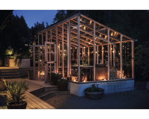 Gewächshaus mit Holzrahmen, Glaswänden und Beleuchtung im Garten bei Nacht.