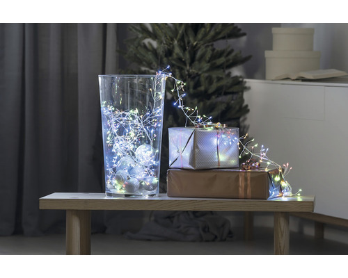 Dekorative Szene mit Glasvase, Lichterkette und Geschenken auf einem Tisch vor einem Weihnachtsbaum