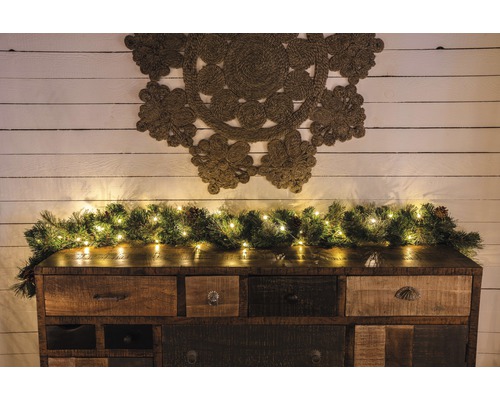 Tropfenlichterkette Konstsmide Lichtfarbe bernstein 50 LEDs Dekoratives Sideboard mit Tannengirlande und Lichterkette sowie einem Wandteppich an der Wand.
