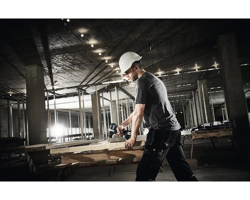 Handwerker mit Schutzhelm bohrt mit einer Bohrmaschine in ein Holzbrett auf einer Baustelle