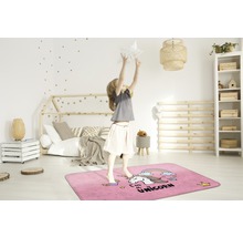 Ein Mädchen springt auf einem rechteckigen, rosafarbenen Teppich mit Einhornmotiv in einem Kinderzimmer.