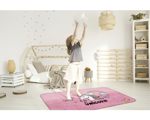 Ein Mädchen springt auf einem rechteckigen, rosafarbenen Teppich mit Einhornmotiv in einem Kinderzimmer.