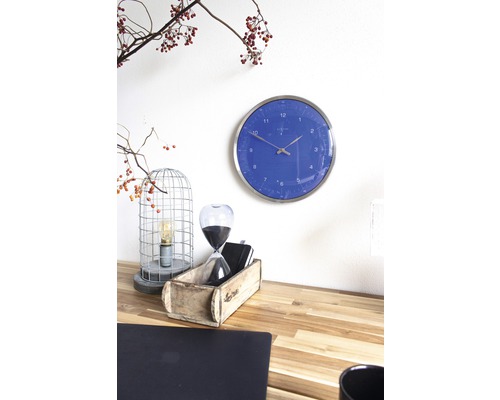 Szene mit einer blauen Wanduhr, einer Sanduhr und einer dekorativen Lampe auf einem Schreibtisch.