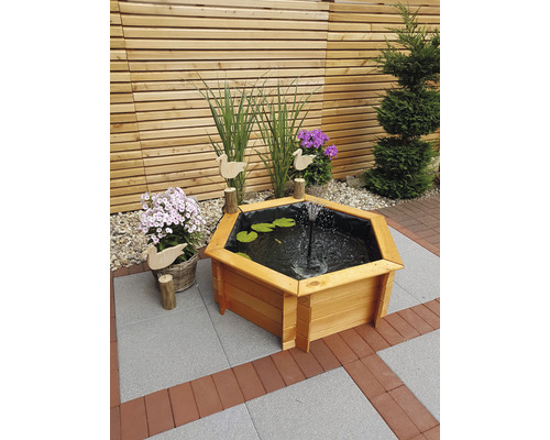 Hexagonaler Holzbrunnen mit Seerosenblättern und einer Fontäne im Garten.