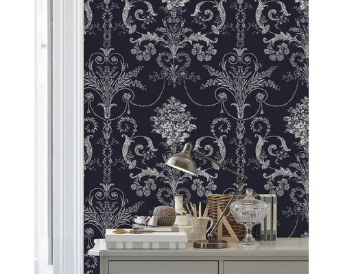 Nahaufnahme eines Raumes mit einer Wand, die mit einer gemusterten Tapete bedeckt ist. Auf einem Tisch stehen verschiedene dekorative Gegenstände und eine Lampe.