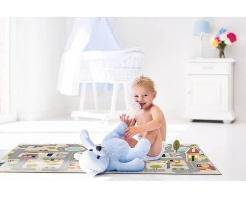 Ein Kind sitzt in einem hellen Raum auf einer Spielmatte mit Straßenmuster und trinkt aus einer Flasche. Neben ihm liegt ein Plüschbär.