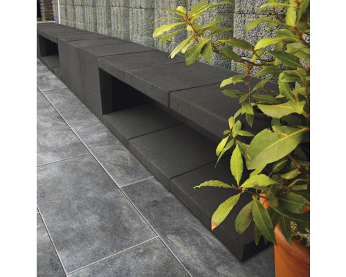 Lange, dunkle Gartenbank aus Betonsteinen neben einem grünen Strauch und Fliesenboden.