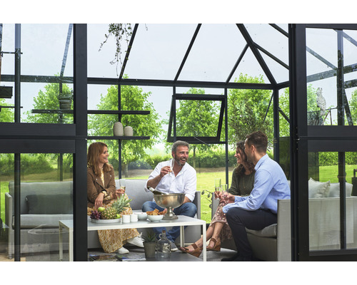 Gruppe von Personen genießt eine gesellige Runde in einem Gewächshaus mit Sitzgelegenheiten und Tisch.