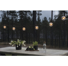 Szene mit einer Lichterkette, Blumen und Getränken auf einem Tisch im Freien vor einem Wald