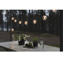 Beleuchtete Lichterkette über einem Tisch im Außenbereich mit Blumen, Pflanzen und Getränken