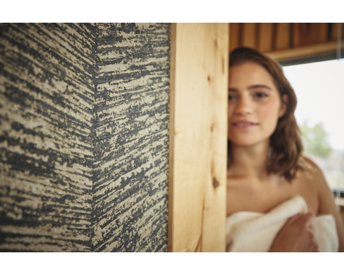 Nahaufnahme einer mit einem Handtuch bedeckten Frau neben einer strukturierten Wand in einem Raum mit Holzakzenten.