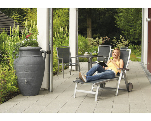 Szene mit einer Frau, die auf einer Liege entspannt, einem Regentank in Amphorenform und Gartenmöbeln auf einer Terrasse.