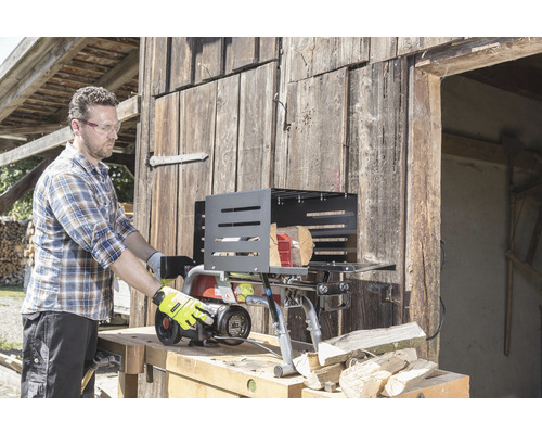 Mann bedient einen Holzspalter vor einer Holzwand. Der Holzspalter steht auf einer Werkbank und neben ihm liegen Holzscheite.