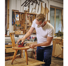 Mann schleift einen Holzstuhl mit einem Exzenterschleifer in einer Werkstatt.