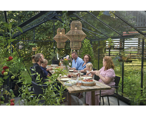 Szene in einem Gewächshaus mit einer Familie, die an einem Holztisch sitzt, umgeben von Pflanzen und zwei Hängelampen.