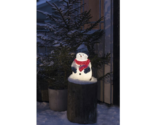 Leuchtender Schneemann mit Mütze und Schal auf einem Baumstumpf, umgeben von einem schneebedeckten Tannenbaum.