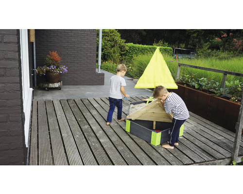 Zwei Kinder spielen in einem Sandkasten mit Dach auf einer Holzterrasse.