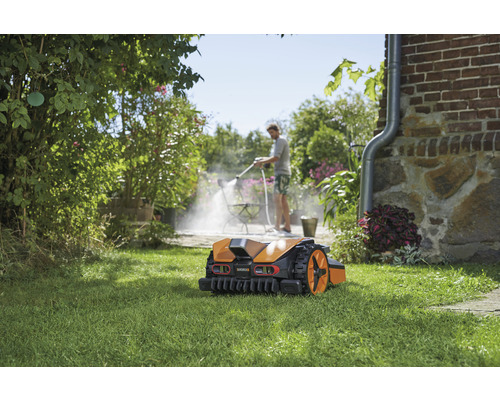 Rasenmähroboter auf Rasen, im Hintergrund reinigt eine Person eine Terrasse mit einem Hochdruckreiniger