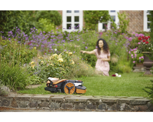 Ein Rasenmähroboter steht auf dem Rasen vor einem blühenden Garten mit einer Frau im Hintergrund.