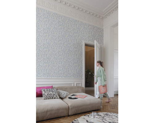 Heller Raum mit floral gemusterter Tapete, Sofa und einer jungen Frau, die durch eine offene Tür geht