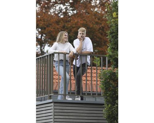 Zwei Personen auf einem Balkon mit Metallgeländer vor einem Haus mit Ziegeldach.