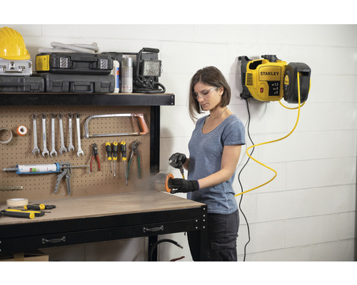 Frau arbeitet mit einem Druckluftkompressor von Stanley in einer Werkstatt. Verschiedene Werkzeuge sind an einer Werkbank befestigt.